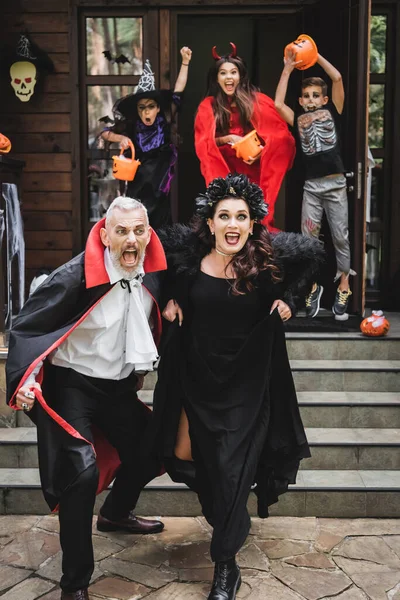 Asustada pareja en halloween trajes corriendo fuera casa de espeluznantes niños - foto de stock