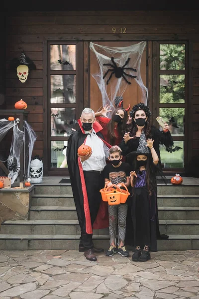 Família assustadora em trajes de Halloween e máscaras médicas pretas mostrando gestos assustadores perto do alpendre com decoração — Fotografia de Stock