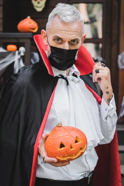 Чоловік середнього віку в чорній медичній масці і костюм вампіра Хеллоуїн тримає різьблений гарбуз, дивлячись на камеру — стокове фото