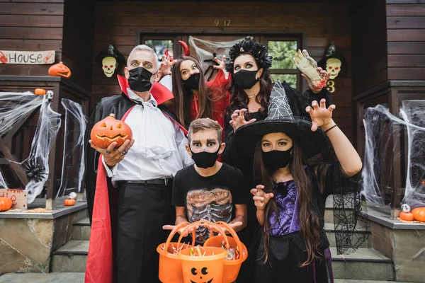 Familia en trajes de Halloween y máscaras médicas negras de pie con cubos de dulces cerca de la casa - foto de stock