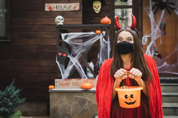 Chica en traje de diablo y máscara médica negro de pie con cubo de dulces cerca de cabaña borrosa con decoración de halloween - foto de stock