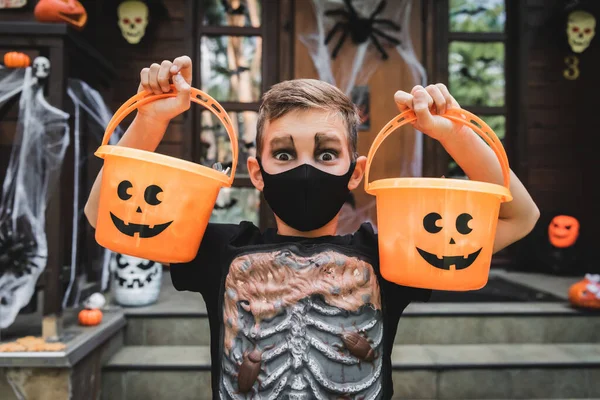 Ragazzo in costume scheletro e maschera medica nera che tiene secchi di Halloween vicino al portico del cottage sfocato — Foto stock