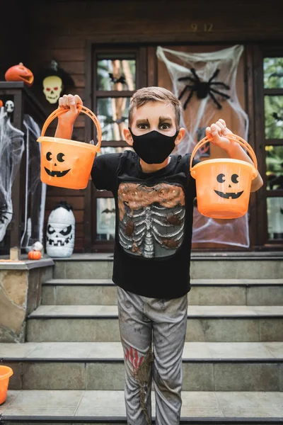 Garçon en costume squelette et masque médical noir tenant seaux halloween près flou chalet — Photo de stock