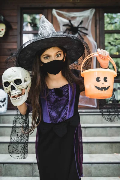 Chica en traje de Halloween bruja y máscara médica negro celebración de cráneo y cubo cerca de casa borrosa - foto de stock