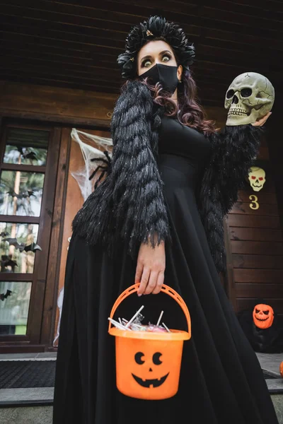 Mujer en traje de Halloween vampiro y máscara médica negro, la celebración de cráneo y cubo de dulces en el porche de la casa - foto de stock