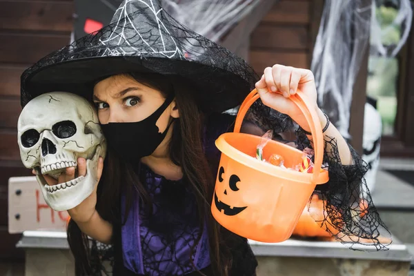 Enfant en chapeau de sorcière et masque médical noir tenant le crâne et le seau avec des bonbons tout en regardant la caméra — Photo de stock