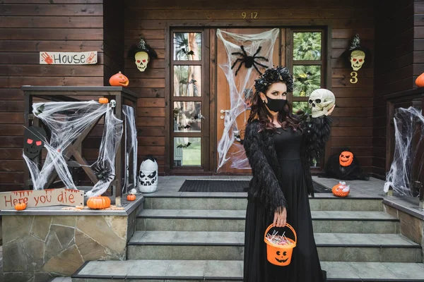 Frau in Halloween-Kostüm und schwarzer medizinischer Maske steht mit Totenkopf und Eimer voller Süßigkeiten in der Nähe der geschmückten Veranda — Stockfoto