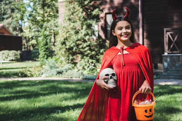 Счастливое дитя в красном костюме демона стоящее с черепом и ведром конфет на улице — стоковое фото