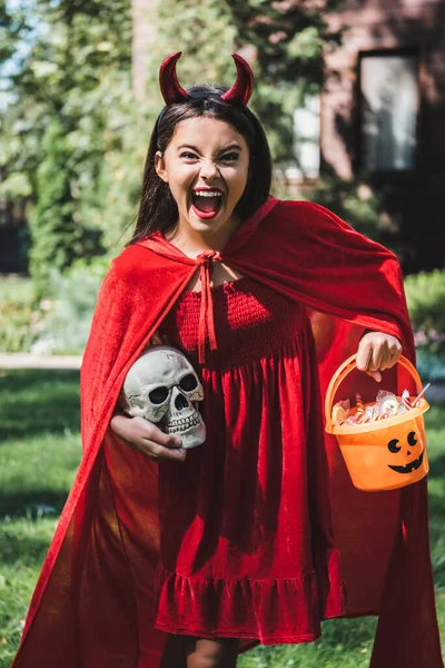 Chica emocionada en disfraz de Halloween diablo gritando mientras sostiene cráneo y cubo de caramelos - foto de stock
