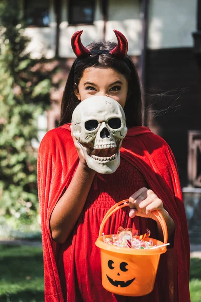 Alegre niño en diablo halloween traje oscuro cara con espeluznante cráneo mientras sostiene cubo de dulces - foto de stock