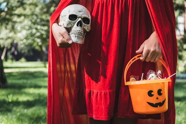 Vue partielle de fille portant costume rouge d'Halloween et tenant crâne et seau de bonbons — Photo de stock