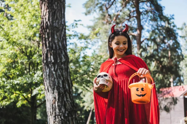 Усміхнена дитина в костюмі диявола Хелловін тримає череп і відро з цукерками в лісі — стокове фото