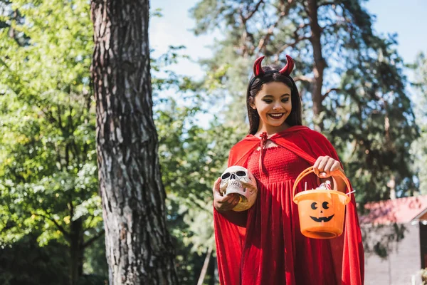 Lächelndes Kind im Teufelskostüm mit Totenkopf und Eimer mit Bonbons im Wald — Stockfoto