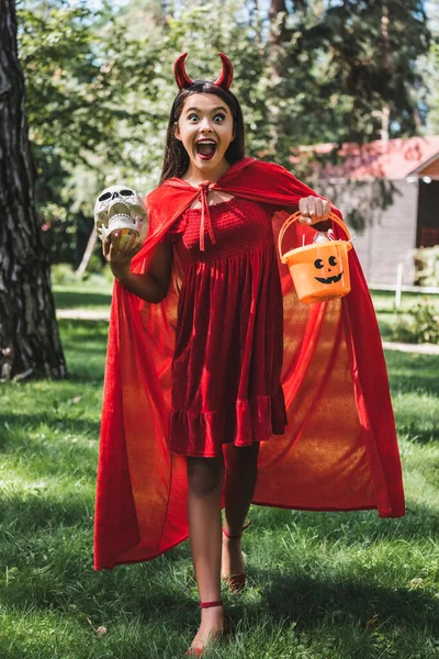 Дивовижна дівчина в костюмі демона Хеллоуїн кричить, стоячи з черепом і відром цукерок на відкритому повітрі — стокове фото
