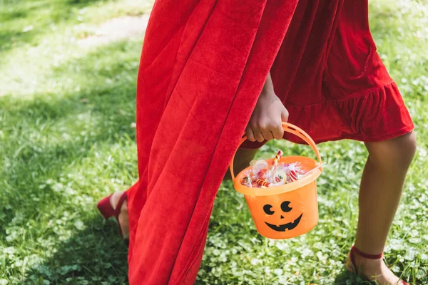 Cropped view of girl in red costume carrying halloween bucket with candies while walking on grass — Stock Photo