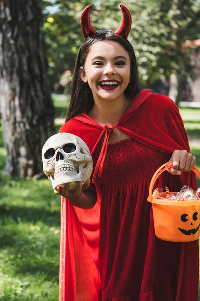 Cheerful girl in demon halloween costume holding skull and bucket with candies outdoors — Stock Photo