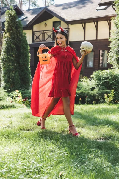 Allegra ragazza in costume diavolo Halloween a piedi con teschio e secchio di dolci vicino casetta sfocata — Foto stock