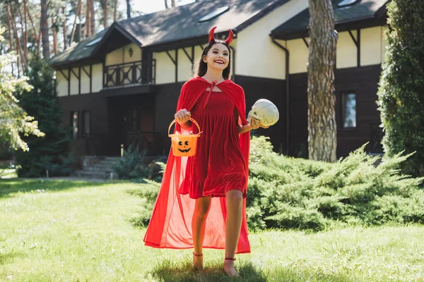 Gioiosa ragazza in costume demone Halloween con teschio e secchio con caramelle vicino alla casa sfocata — Foto stock