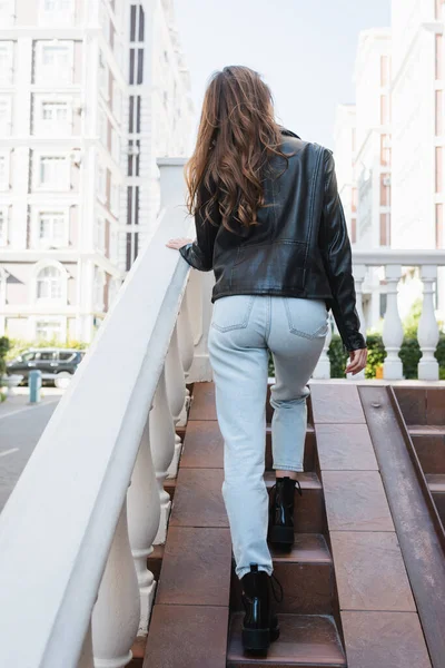 Back view of curly young woman in black leather jacket walking on stairs outside — Stock Photo