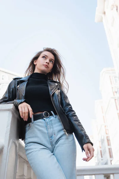 Низький кут зору красивої молодої жінки в чорній шкіряній куртці і джинсах, що стоять на міській вулиці — стокове фото