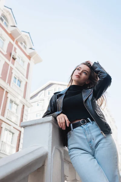 Visão de baixo ângulo da jovem mulher em jaqueta de couro preto e jeans ajustando o cabelo na rua urbana — Fotografia de Stock