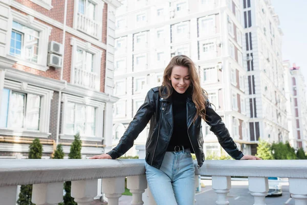 Mujer joven y complacida en chaqueta de cuero negro sonriendo en la calle urbana en Europa - foto de stock