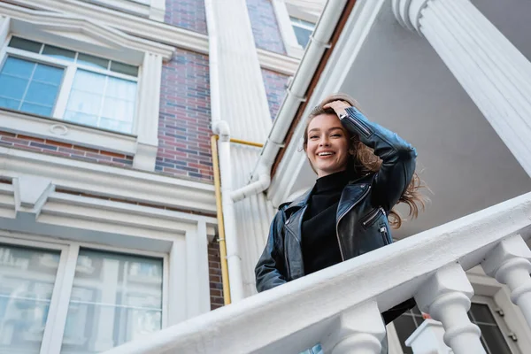 Vue à angle bas de la jeune femme positive en veste en cuir souriant sur la rue urbaine en Europe — Photo de stock