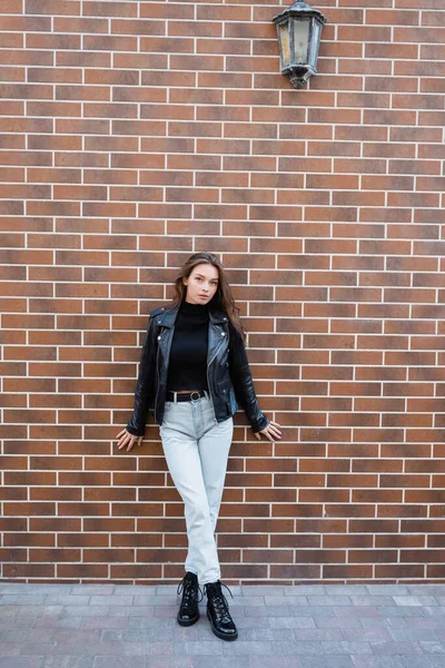 Pleine longueur de jolie jeune femme en veste en cuir et jeans debout près du mur de briques — Photo de stock