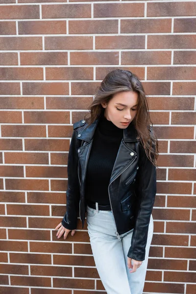 Jolie jeune femme en veste et jeans en cuir debout près du mur de briques — Photo de stock