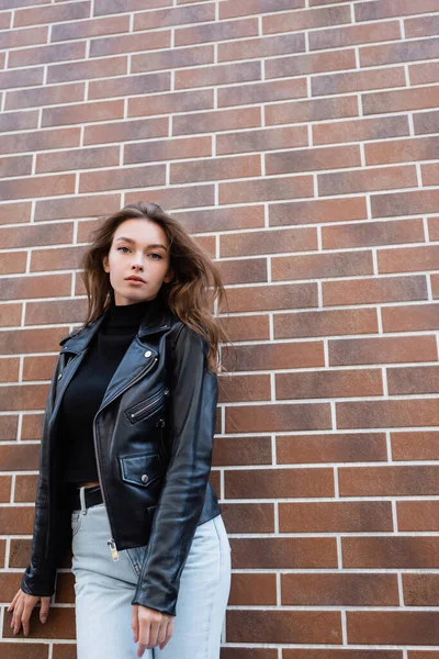 Pretty young woman in black leather jacket and jeans standing near brick wall — Stock Photo