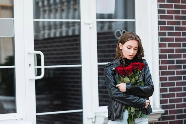 Jovem mulher em jaqueta de couro olhando para rosas vermelhas na rua urbana na europa — Fotografia de Stock