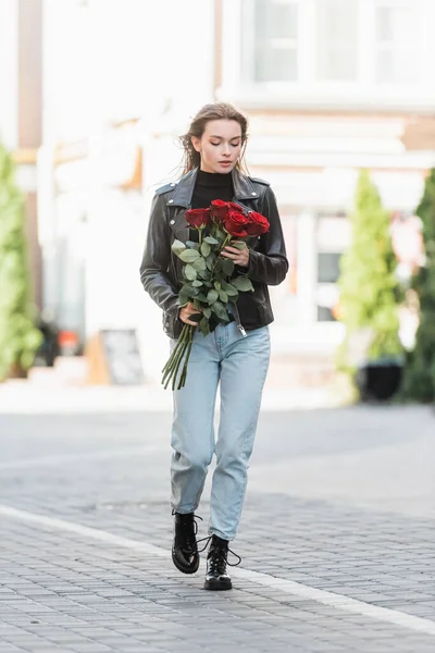 Comprimento total da mulher bonita em jaqueta de couro segurando rosas vermelhas e andando na rua urbana na europa — Fotografia de Stock