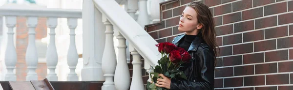 Bonita mujer en chaqueta de cuero que sostiene ramo de rosas rojas en la calle urbana en Europa, pancarta - foto de stock