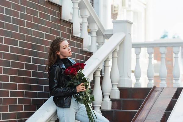 Pretty woman in leather jacket and jeans holding bouquet of red roses on urban street in europe — Stock Photo