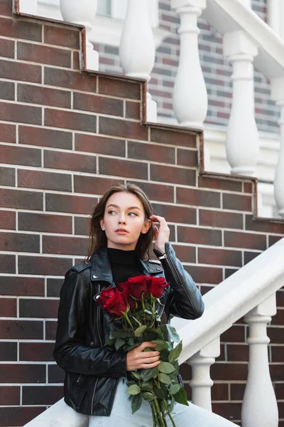 Pretty young woman in leather jacket holding bouquet of red roses outside — Stock Photo