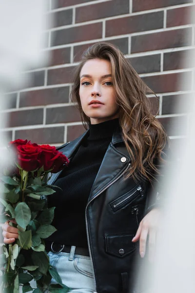 Jolie femme en veste en cuir tenant bouquet de roses rouges et regardant la caméra à l'extérieur — Photo de stock