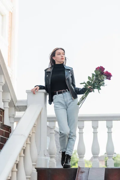 Comprimento total de mulher bonita em jaqueta de couro preto e jeans segurando buquê de rosas na rua urbana na europa — Fotografia de Stock