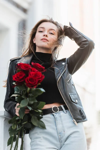 Pretty woman in black leather jacket and jeans holding bouquet of roses on urban street — Stock Photo