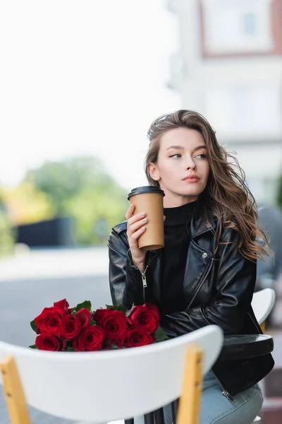 Bonita mujer en chaqueta de cuero negro sosteniendo taza de papel cerca de ramo de rosas en la mesa de café - foto de stock