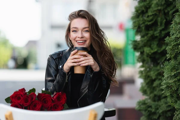 Cheerful woman in leather jacket holding paper cup near bouquet of red roses on coffee table — Stock Photo