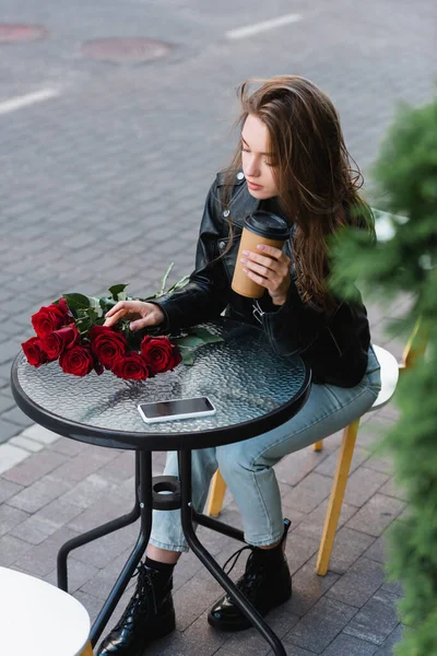 High angle view of pretty woman in leather jacket holding paper cup near bouquet of red roses and smartphone on coffee table — Stock Photo