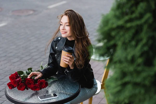 High angle view of pleased woman in leather jacket holding paper cup near smartphone and bouquet of red roses on coffee table — Stock Photo