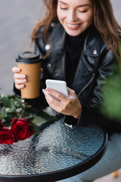 Cheerful blurred woman in leather jacket holding paper cup and using smartphone near bouquet of red roses — Stock Photo