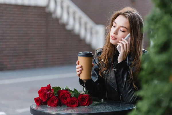 Hübsche Frau in Lederjacke hält Pappbecher in der Hand und spricht auf dem Smartphone neben einem Strauß roter Rosen — Stockfoto