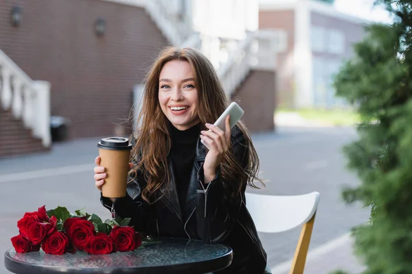 Cheerful woman in leather jacket holding paper cup and smartphone near bouquet of red roses — Stock Photo