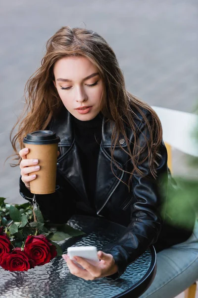 Jeune femme veste en cuir tenant tasse en papier et smartphone près bouquet de roses rouges — Photo de stock