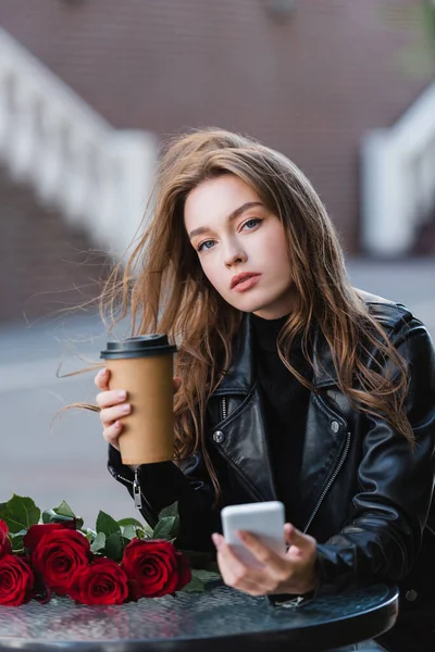 Mujer joven en chaqueta de cuero sosteniendo café para llevar y teléfono inteligente cerca de ramo de rosas rojas - foto de stock