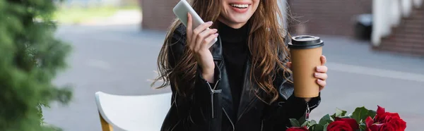 Vista ritagliata di donna allegra in giacca di pelle con tazza di carta e smartphone vicino al mazzo di rose rosse, banner — Foto stock