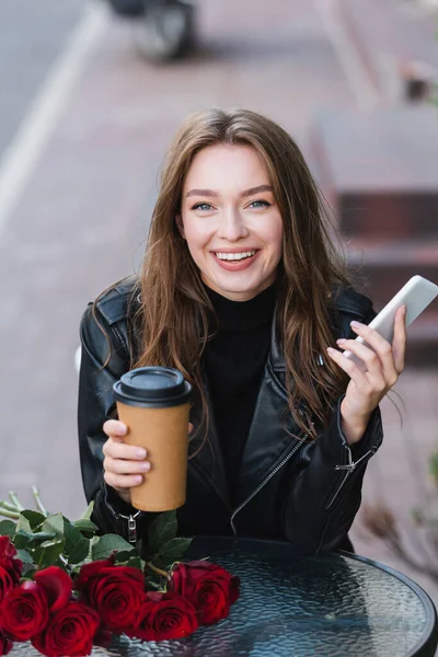 Femme souriante veste en cuir tenant tasse en papier et smartphone près du bouquet de roses rouges — Photo de stock