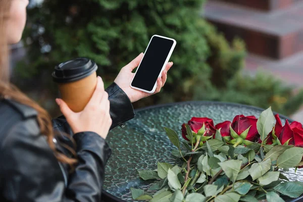 Vista recortada de la mujer joven sosteniendo teléfono inteligente con pantalla en blanco y taza de papel cerca de rosas en la terraza de la cafetería - foto de stock